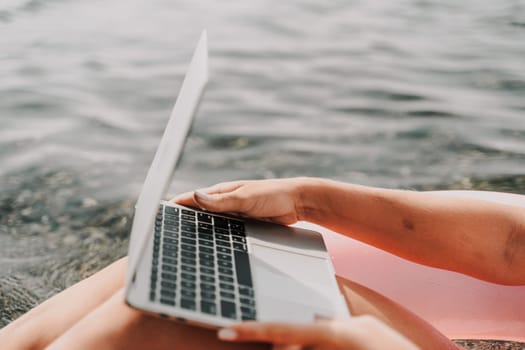 A woman is sitting on the beach with a laptop open in front of her. She is typing on the keyboard and she is working or studying. Concept of relaxation and productivity