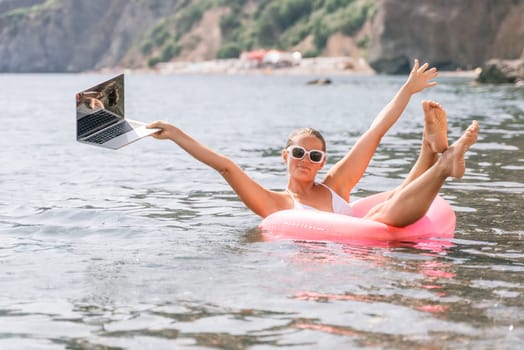 A woman is holding a laptop while floating in a pink inflatable raft on a lake. The scene is playful and fun, with the woman seemingly enjoying her time in the water