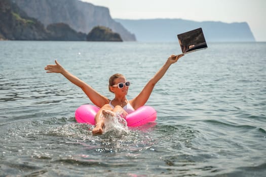 A woman is floating in a pink inflatable raft in the ocean. She is holding a laptop in her hand