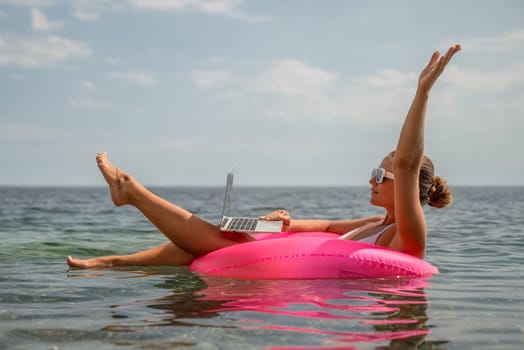 A woman is sitting on a pink inflatable raft in the ocean, using a laptop. Concept of relaxation and leisure, as the woman is enjoying her time in the water while working on her laptop