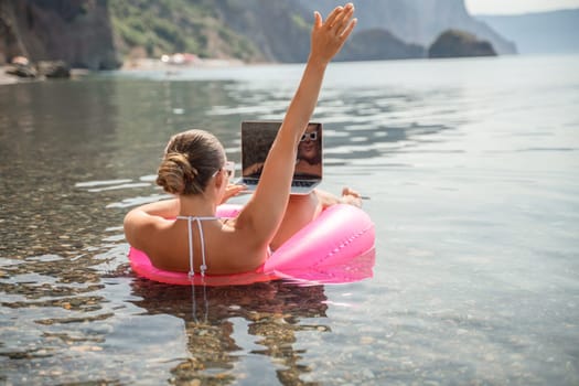 A woman is floating on a pink inflatable raft in the ocean while holding a laptop. Concept of relaxation and leisure, as the woman enjoys her time in the water while working on her laptop