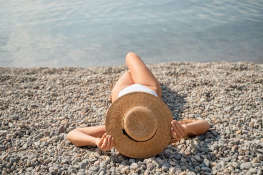 A woman is laying on the beach wearing a straw hat and a white bikini. She is enjoying the sun and the beach