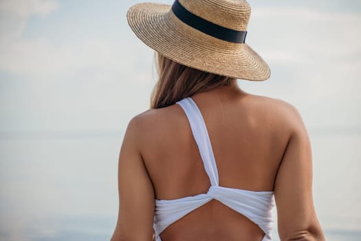 A woman wearing a straw hat and a white bikini top is standing on a beach. The hat is tilted to the side, and the woman's hair is blowing in the wind. The beach is calm