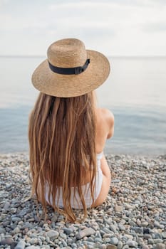 A woman with long hair is sitting on a beach with a straw hat on. The hat is black and white