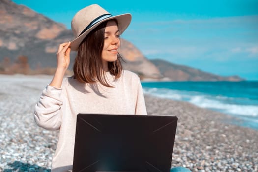 A young smiley girl in a hat and casual sweater sits on the beach by the sea with a laptop on a sunny day, works, studies, buys tickets during trip, woman rests on vacation and types on the keyboard.