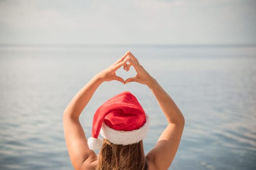 A woman wearing a red Santa hat is holding her hands together in a heart shape over the water