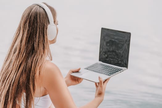 A woman is sitting by the water with a laptop in her lap. She is wearing headphones and she is focused on her work. Concept of productivity and relaxation