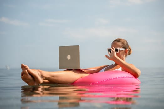 A woman is sitting on a pink inflatable raft in the water, using a laptop. Concept of relaxation and leisure, as the woman is enjoying her time in the water while working on her laptop