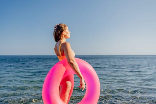 A woman is standing on the beach holding a pink inflatable ring. The beach is calm and the sky is clear