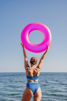 A woman is holding a pink inflatable ring in the air. The ring is shaped like an o. The woman is standing on the beach, and the ocean is in the background. The scene is bright and cheerful