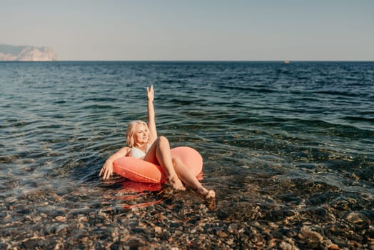A woman is floating in a red inflatable raft on a rocky beach. She is holding her hands up in the air, possibly making a peace sign. The scene is peaceful and relaxing
