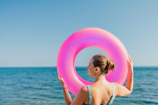 A woman is holding a pink inflatable ring in the ocean. The scene is bright and cheerful, with the woman's hair pulled back and the water sparkling in the sunlight