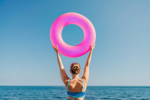 A woman is holding a pink inflatable ring above the ocean. Concept of fun and relaxation, as the woman is enjoying her time at the beach. The pink color of the ring adds a playful