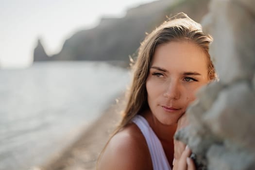 A woman with long hair is looking out at the ocean. She is wearing a white tank top and she is lost in thought