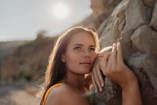 A woman in a yellow tank top is leaning against a rock. She has a smile on her face and her eyes are closed