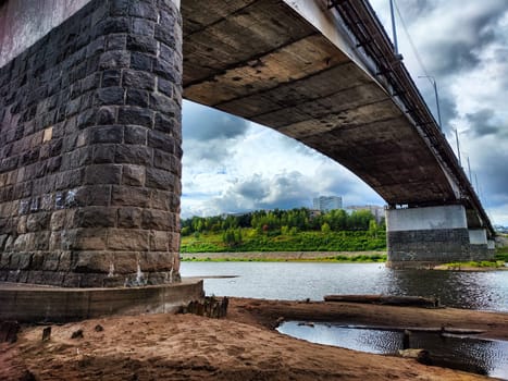 A large bridge extends over a calm river, with an overcast sky and urban buildings visible in the background