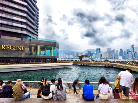 Hong Kong - April 05, 2024: People gather on the Hong Kong waterfront, with skyscrapers and Victoria Bay in the background on a cloudy afternoon