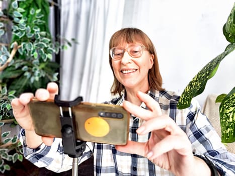 Smiling woman in glasses using a smartphone on a tripod for a video chat. A middle-aged woman posing and taking a selfie