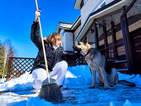 A girl or a woman with a snow broom at home and a large German Shepherd dog. Woman Clearing Snow at Home With Her German Shepherd