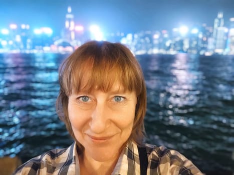 A middle-aged woman smiles for selfie with the Victoria Bay and Hong Kong skyline in the background at night