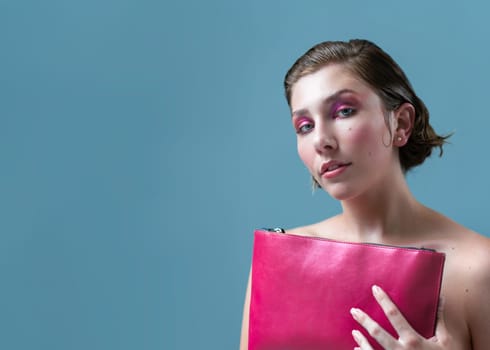 Portrait of topless young female model with short brown hair and eyeshadow, holding pink purse and looking directly at camera over blue background with empty mockup space.