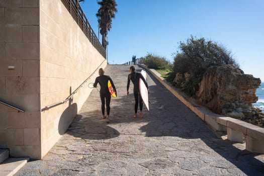 Full body of anonymous women in wetsuit carrying surfboards while walking on footpath near sea. Female friends leaving beach after surfboarding under blue sky on sunny day.