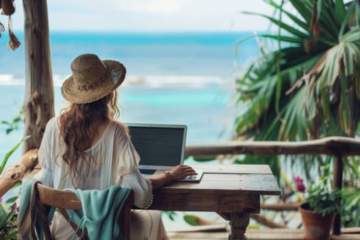 Woman in straw hat working on laptop at wooden table overlooking ocean beach travel and business concept