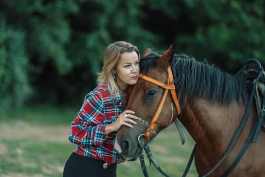 Happy blonde with horse in forest. Woman and a horse walking through the field during the day. Dressed in a plaid shirt and black leggings