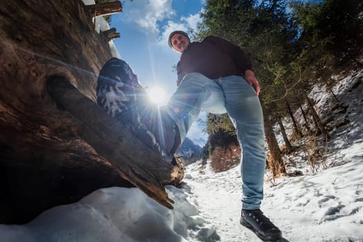 Funny tourist stands with one foot on a fallen tree while traveling along a hiking winter mountain trail.