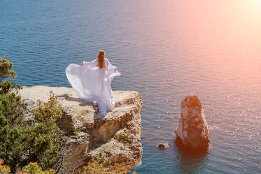 Woman in a white dress on the sea. Side view Young beautiful sensual woman in white long dress posing on a rock high above the sea at sunset. Girl in nature against the blue sky.