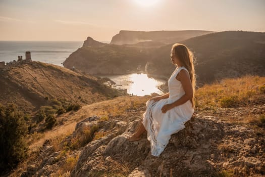 A woman in a white dress sits on a rock overlooking a body of water. The scene is serene and peaceful, with the woman enjoying the view and the calmness of the surroundings