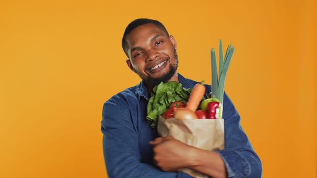 Male model hugging his paper bag full of organic homegrown food, enjoying heathy eating from local bio supermarket. Young vegan person recommending ethically sourced produce. Camera A.