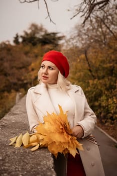 autumn woman in a red beret, a light coat and a red skirt, against the backdrop of an autumn park with yellow leaves in her hands.