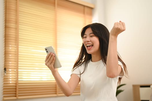 Excited young asian woman reading good news on mobile phone celebrating with yes gesture.