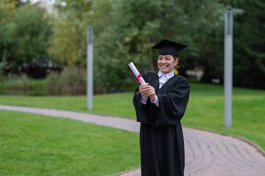 Portrait of happy caucasian woman in graduate gown holding diploma outdoors