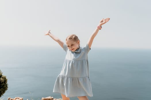 A young girl is standing on a beach, wearing a blue dress and holding her arms up in the air. Concept of joy and freedom, as the girl appears to be enjoying her time by the ocean