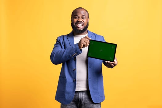 Ecstatic african american man excited while showing green screen tablet, doing recommendation. Jolly BIPOC person delighted to present chroma key portable device, isolated over studio background