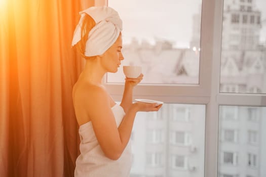 Young serene relaxed woman in spa bath towel drinking hot beverage tea coffee after taking shower bath at home. Beauty treatment, hydration concept