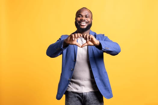 Portrait of loving joyous african american man doing heart symbol shape with hands. Happy caring BIPOC person showing love gesturing, isolated over yellow studio background