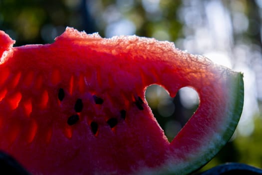 Man holding red watermelon slice cut into heart shape in green garden background. Valentine's Day love concept. Sunset outdoors summer day. Healthy seasonal local food. Traditional