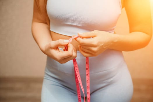 Cropped view of slim woman measuring waist with tape measure at home, close up. European woman checking the result of diet for weight loss or liposuction indoors.