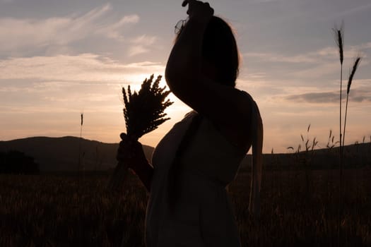 A woman is holding a bunch of wheat in her arms. The wheat is dry and brown, and the woman is wearing a white dress. The scene is set in a field, and the woman is posing for a photo