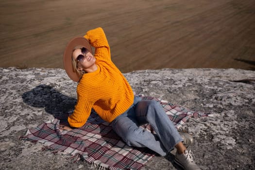 A woman in a yellow sweater and hat is sitting on a blanket in a field. She is smiling and enjoying the outdoors