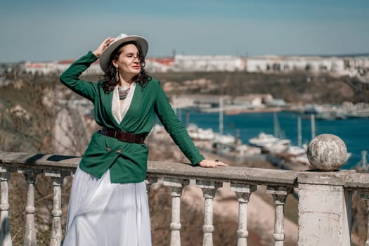 Woman walks around the city, lifestyle. Happy woman in a green jacket, white skirt and hat is sitting on a white fence with balusters overlooking the sea bay and the city