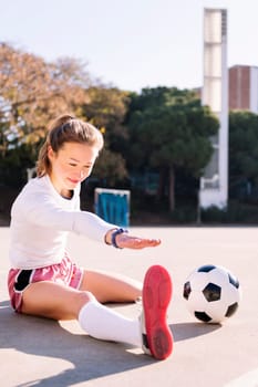 young caucasian woman with a soccer ball stretching legs in a urban football court, concept of sport and active lifestyle, copy space for text