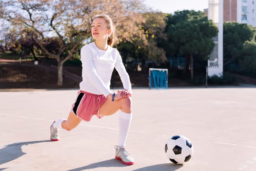 young caucasian woman with a soccer ball stretching legs in a urban football court, concept of sport and active lifestyle