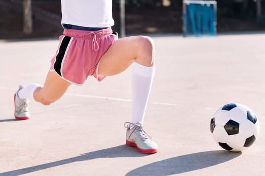 unrecognizable woman with a soccer ball stretching legs in a urban football court, concept of sport and active lifestyle