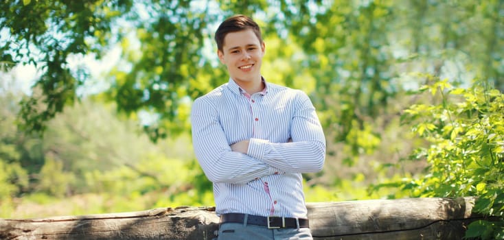 Portrait of handsome happy smiling teenager boy, young guy with crossed arms in shirt standing outdoors in summer park