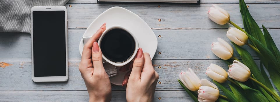 bouquet of white tulips with a Cup of coffee in women's hands, a smartphone and an empty notebook for text. Flat lay, top view