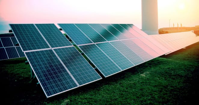 Rows of blue photovoltaic panels stand in an open field, capturing the last light as the sun sets on the horizon. The sky displays a gradient of colors from blue to soft orange, signifying the transition from day to night. The solar farm generates sustainable energy in a rural setting, highlighting the blend of technology with the natural environment.